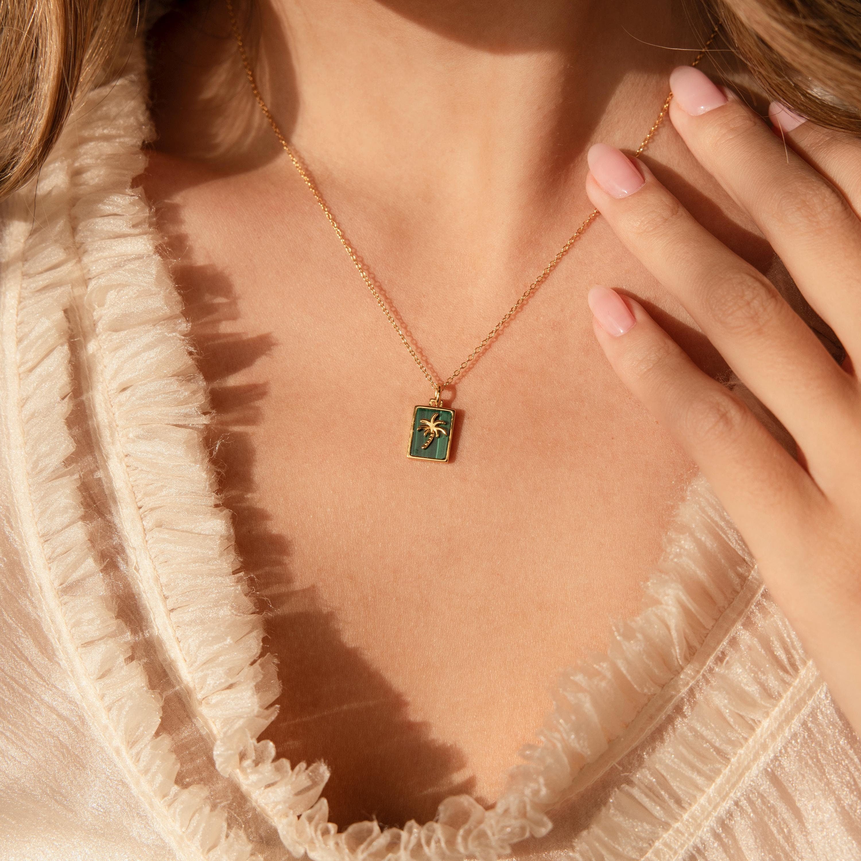 A woman wearing the Malachite Palm Tree Necklace with a gold chain and green rectangular pendant touches it while standing in a beige ruffled top.