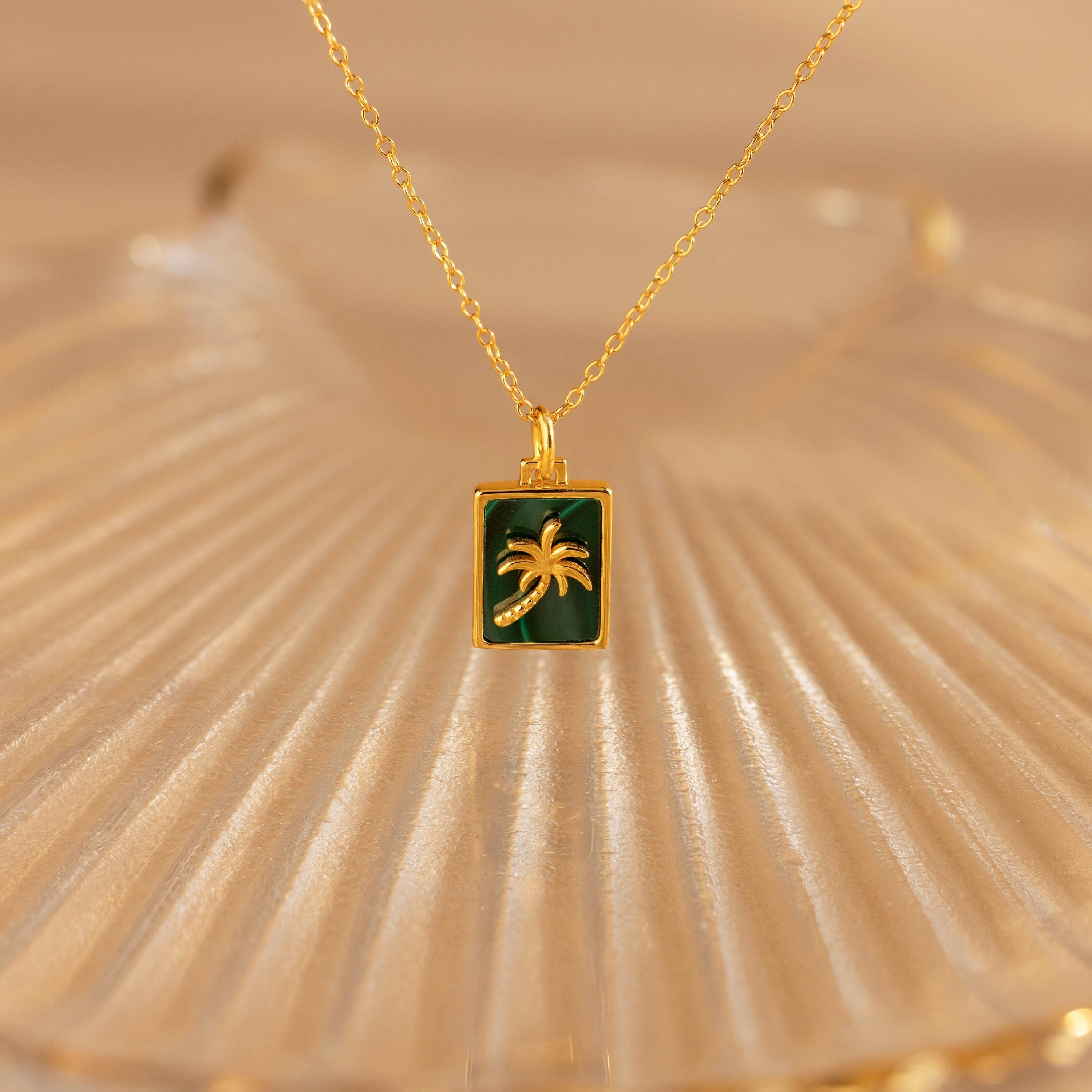 The Malachite Palm Tree Necklace features a rectangular green malachite pendant with a gold palm tree on a gold chain, beautifully displayed on a shell dish—perfect for anyone who loves elegant palm tree jewelry.