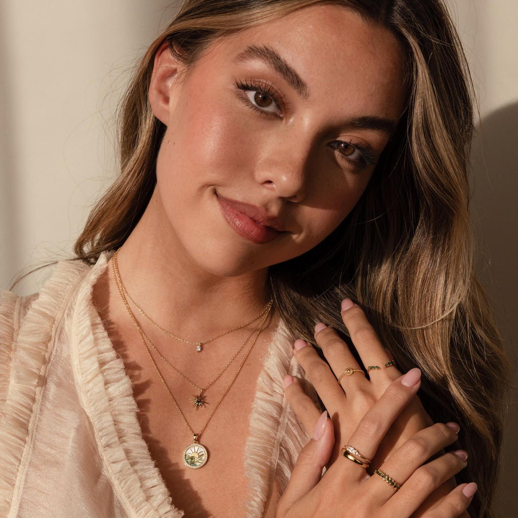 A woman with long hair, smiling softly, wears the Abalone Sunrise Pendant Necklace layered with gold chains, rings, and a sheer ruffled top.