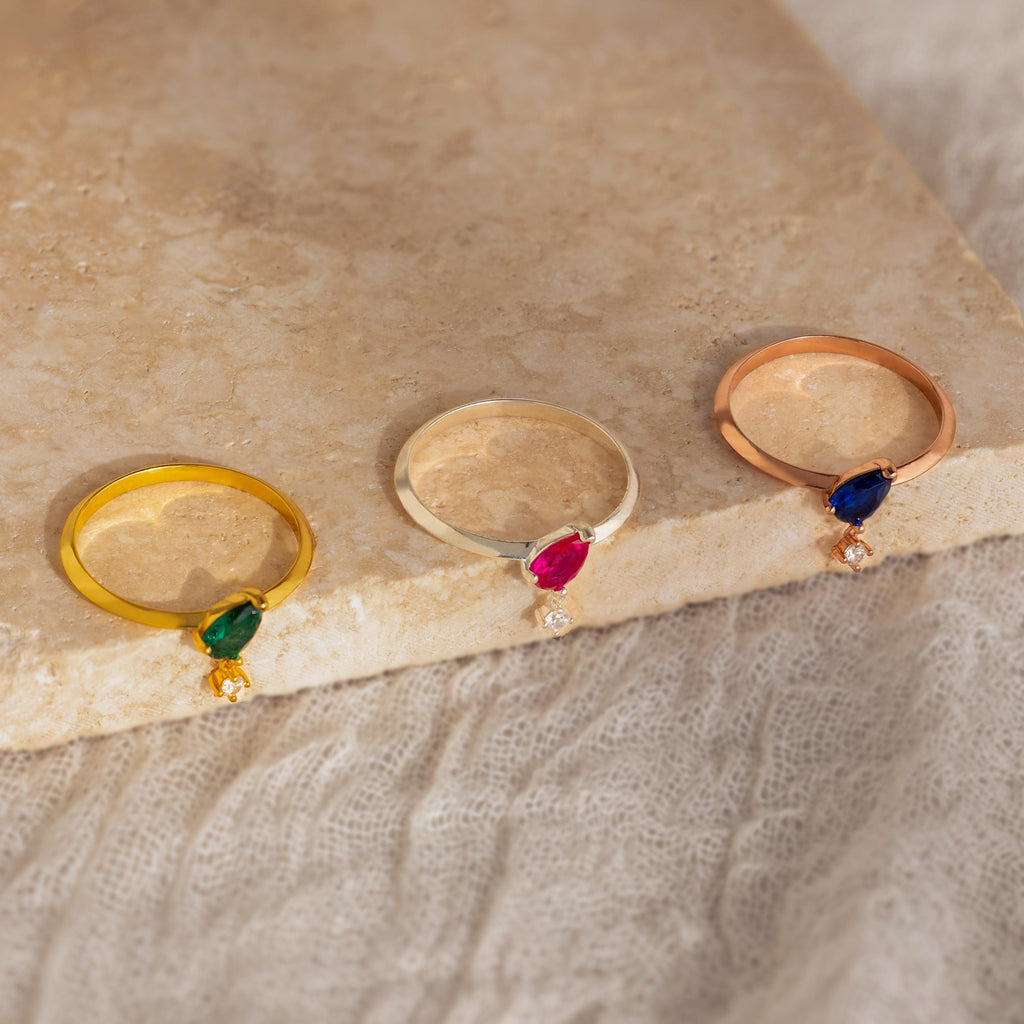 Three rings, featuring the Duo Birthstone Drop Ring and two other customizable designs with small diamonds, are displayed on a stone slab against a textured background.