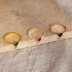 Three rings, featuring the Duo Birthstone Drop Ring and two other customizable designs with small diamonds, are displayed on a stone slab against a textured background.