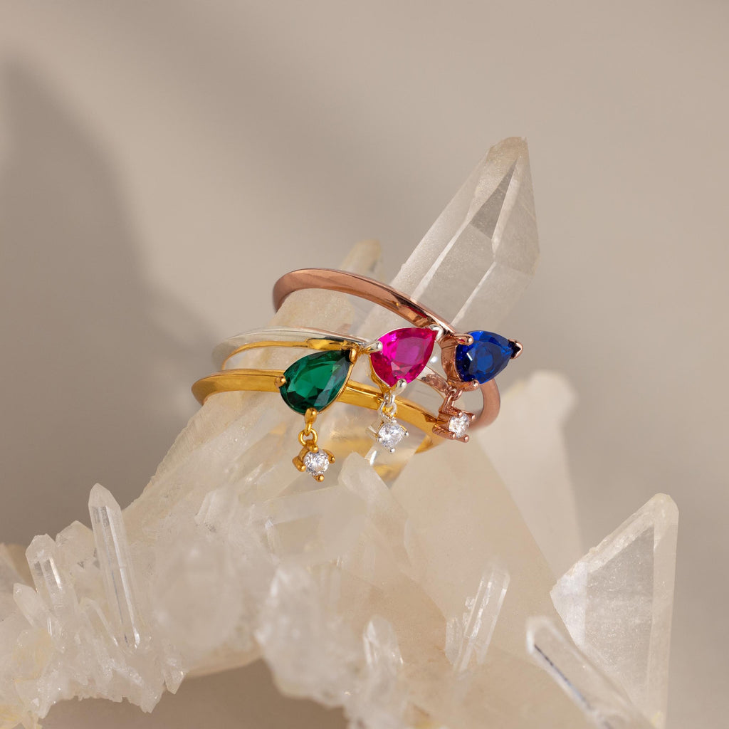 The Duo Birthstone Drop Ring, featuring customizable gemstones, is displayed on clear quartz crystals against a soft, neutral background.