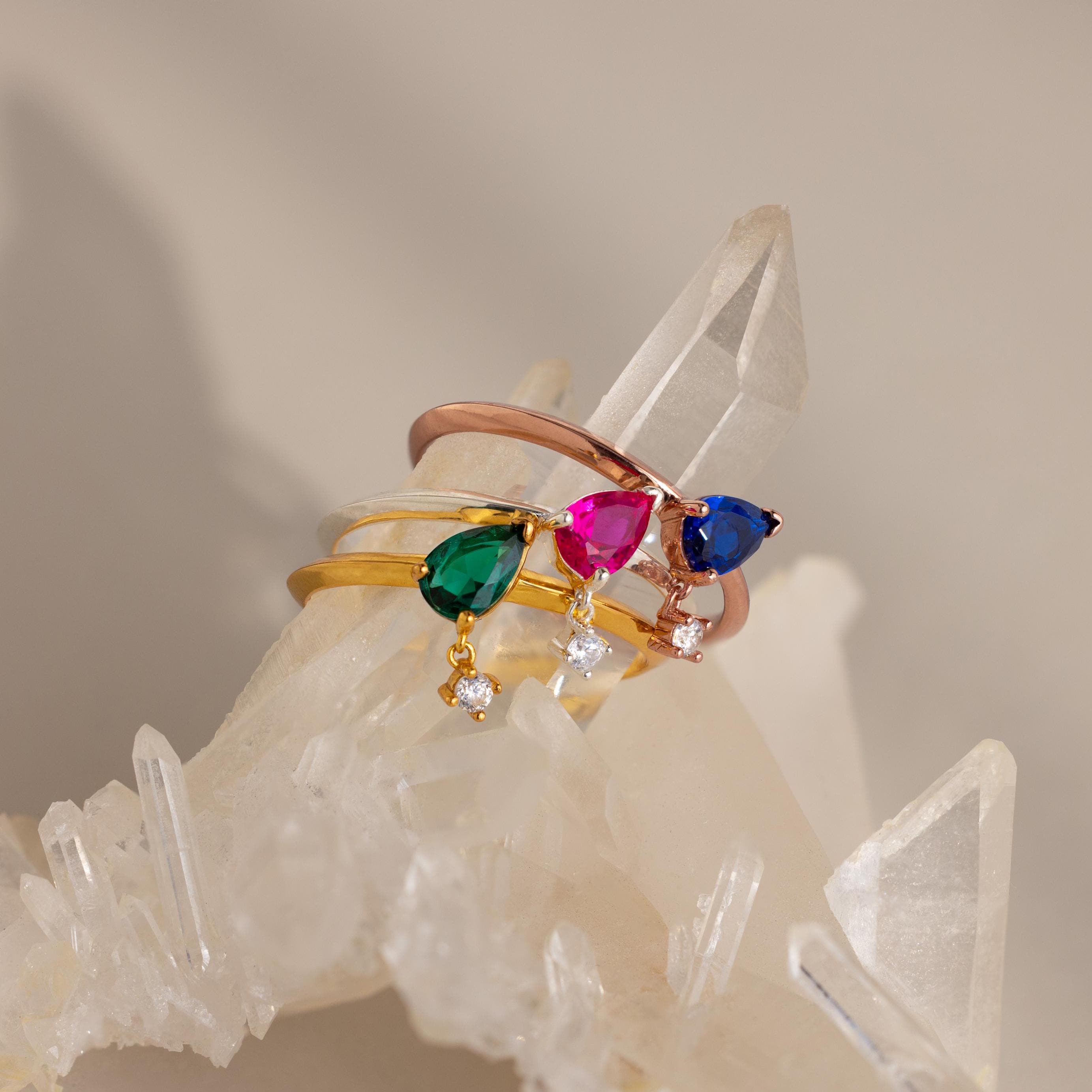 The Duo Birthstone Drop Ring, featuring customizable gemstones, is displayed on clear quartz crystals against a soft, neutral background.