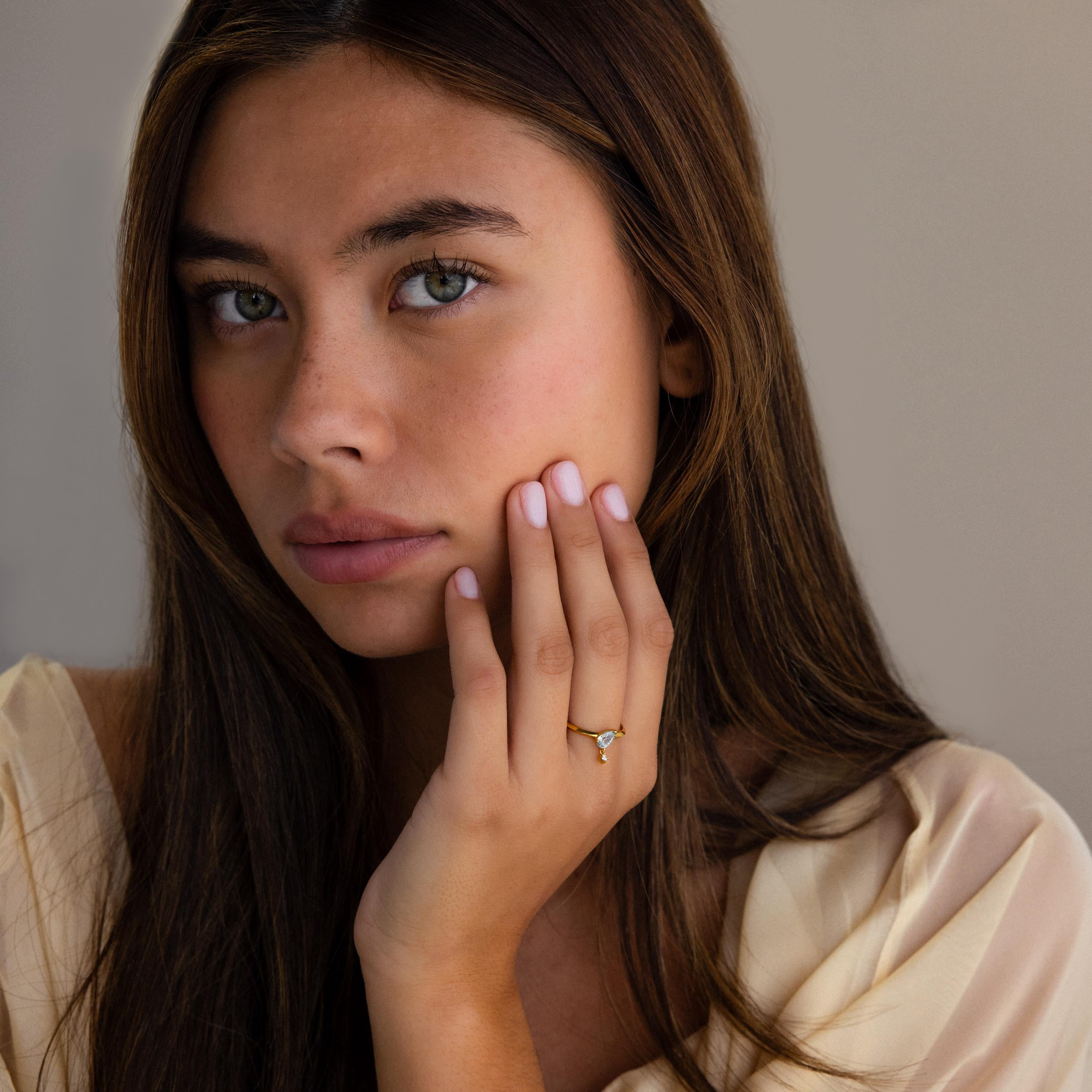 A woman with long brown hair, wearing a cream-colored top and the Duo Birthstone Drop Ring, touches her face while looking at the camera.
