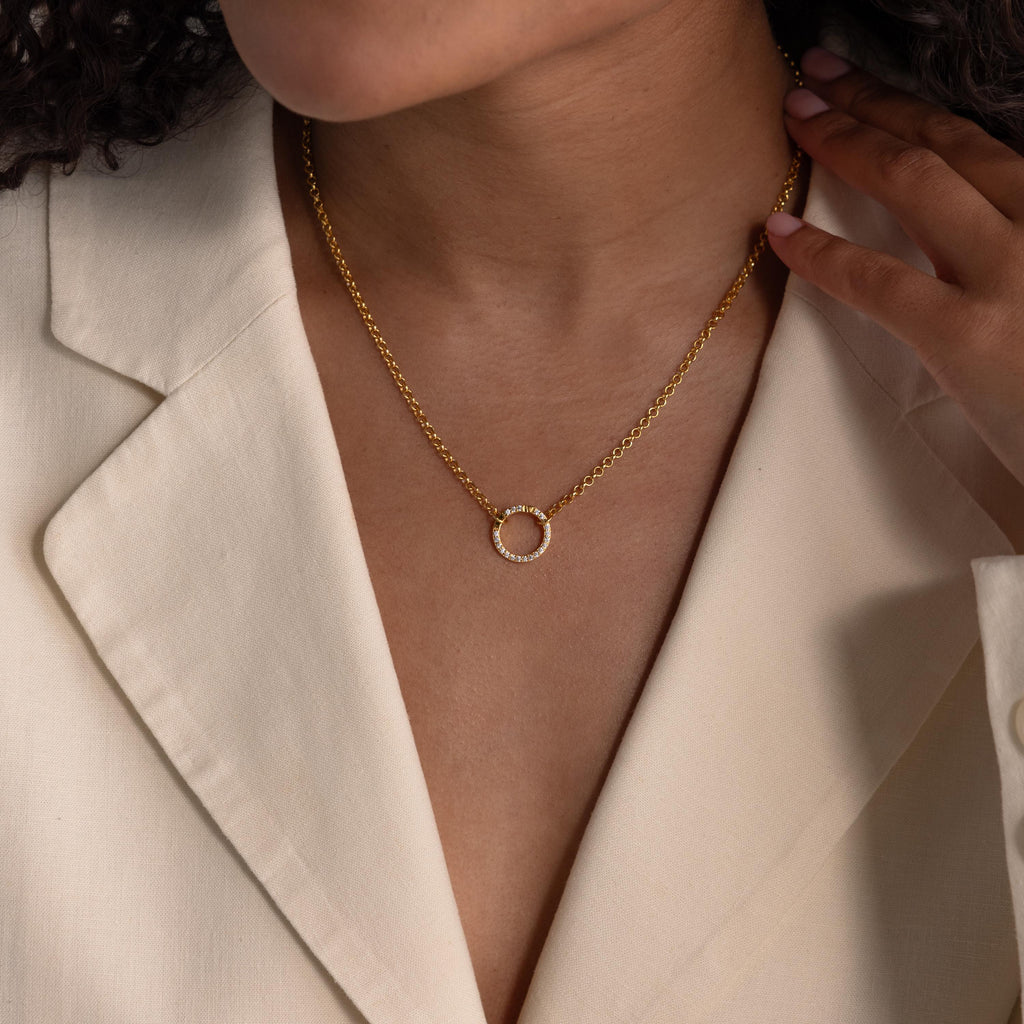 Woman wearing a cream blazer and a gold necklace with a circular Pave Pink Tourmaline Charm Clip pendant, hand touching her collar to showcase this elegant, personalized jewelry piece.