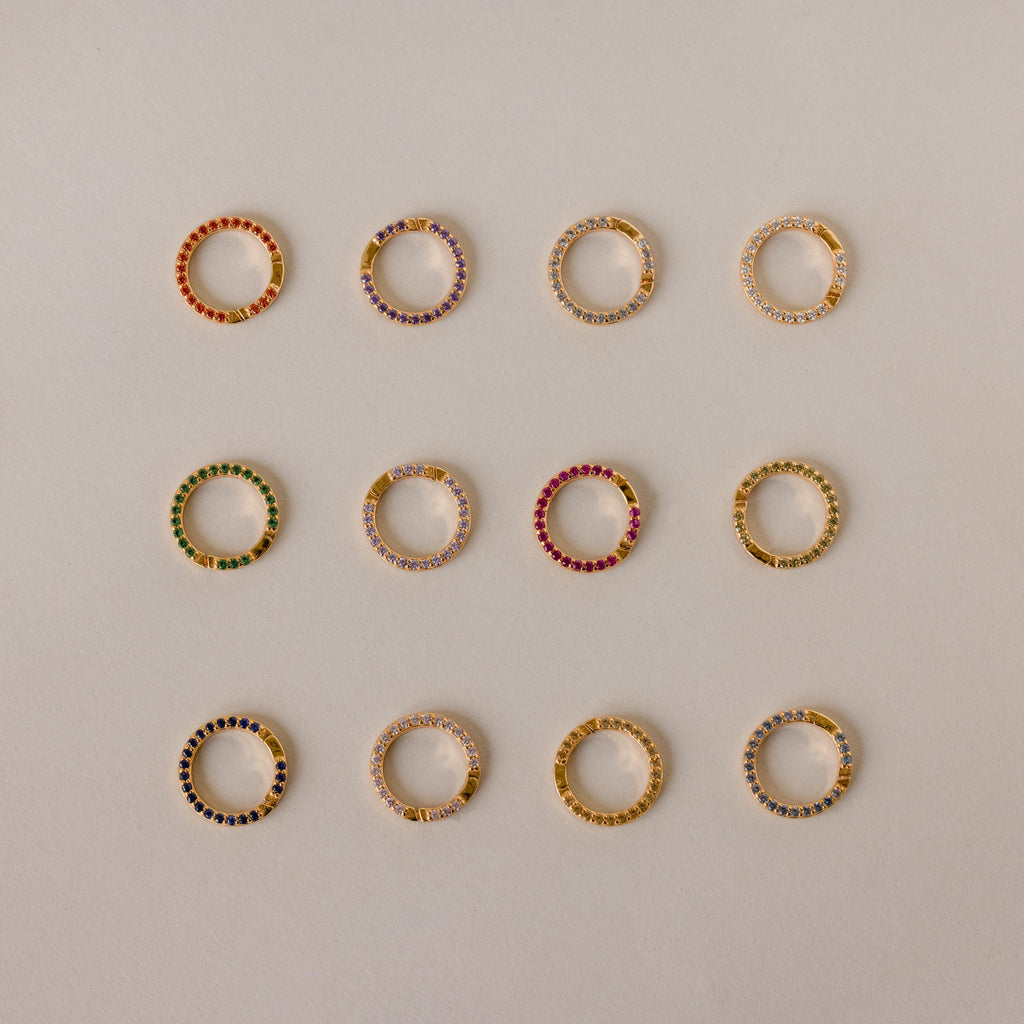 Twelve Pave Birthstone Charm Clips, each a small gold hoop earring with colored stones, arranged in a grid on a beige background—perfect for personalized jewelry.