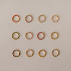 Twelve Pave Birthstone Charm Clips, each a small gold hoop earring with colored stones, arranged in a grid on a beige background—perfect for personalized jewelry.