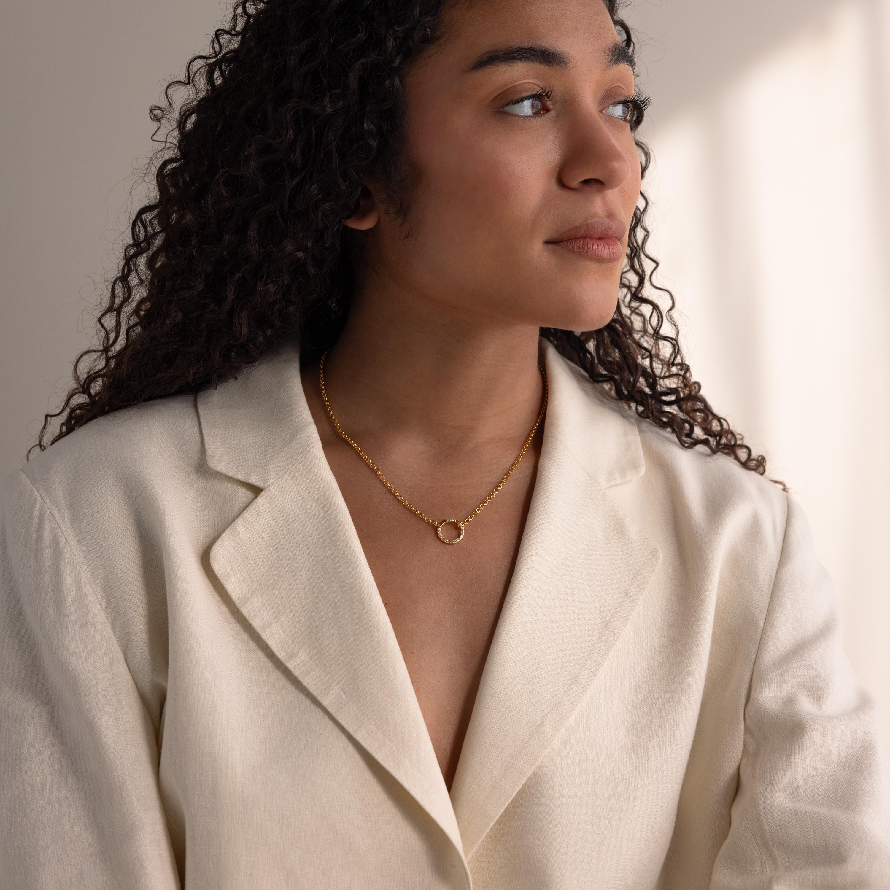 Woman with curly hair wearing a cream blazer and gold necklace, featuring the Pave Diamond Charm Clip as a delicate accent, looking to the side in soft lighting.
