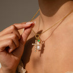 Model wears a gold Rolo Necklace and holds the side up, showcasing dangling gold Pave Cross Charm, long pearl charm, emerald birthstone charm and initial charm  