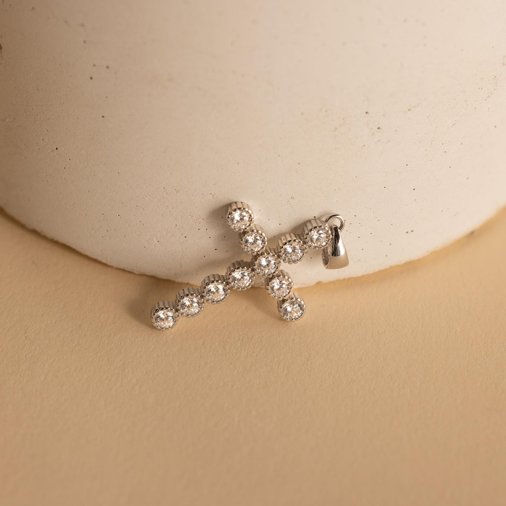 Silver Pave Cross Charm leaning against a vase. The design features multiple round 4mm CZ Diamonds in the shape of a cross with a ribbed bezel setting.