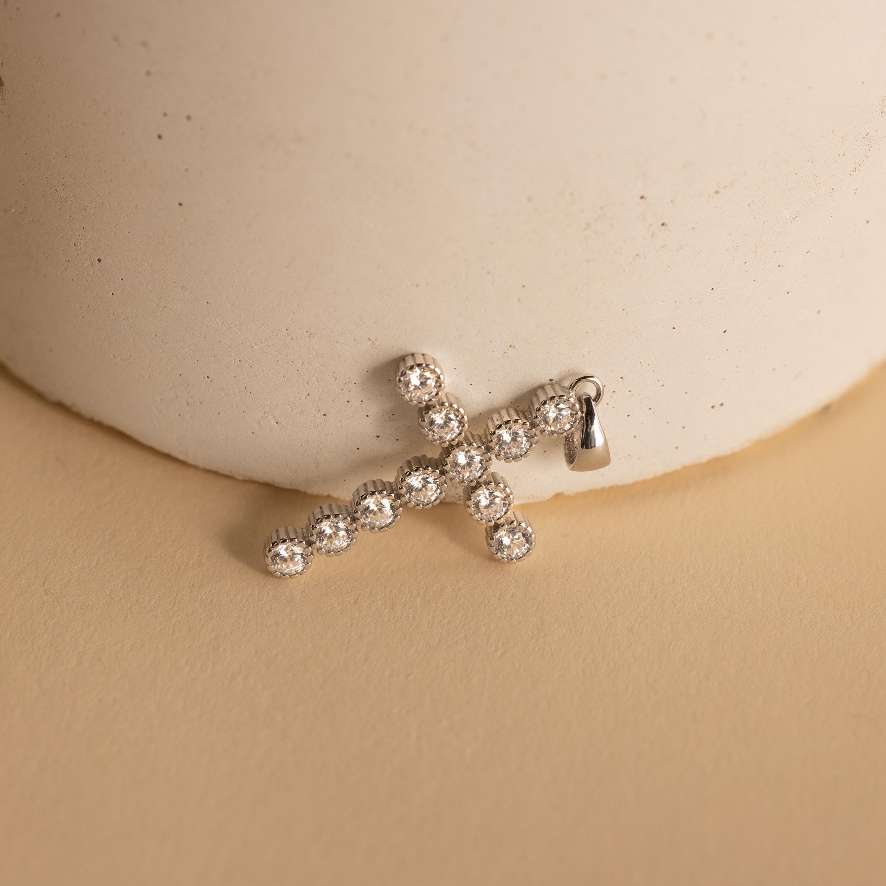 Silver Pave Cross Charm leaning against a vase. The design features multiple round 4mm CZ Diamonds in the shape of a cross with a ribbed bezel setting.