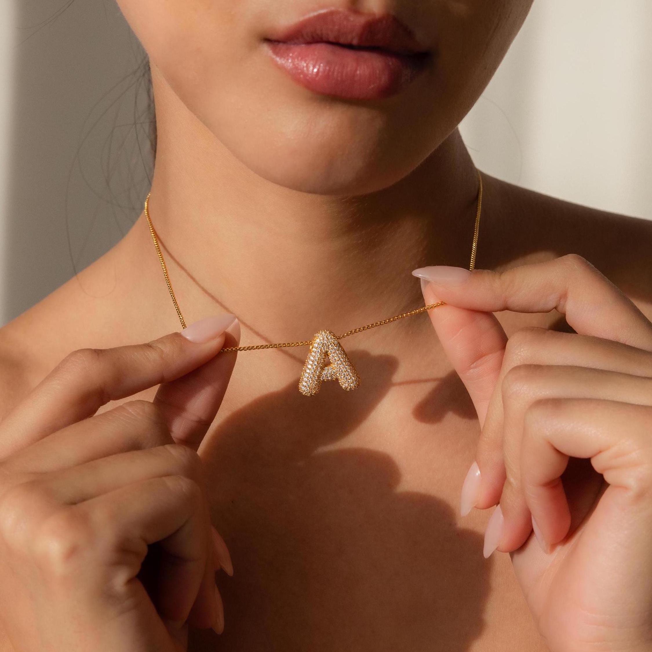 A woman holds the Pave Puff Letter Necklace in 18K Gold, featuring a sparkling pavé diamond letter A pendant, near her collarbone.