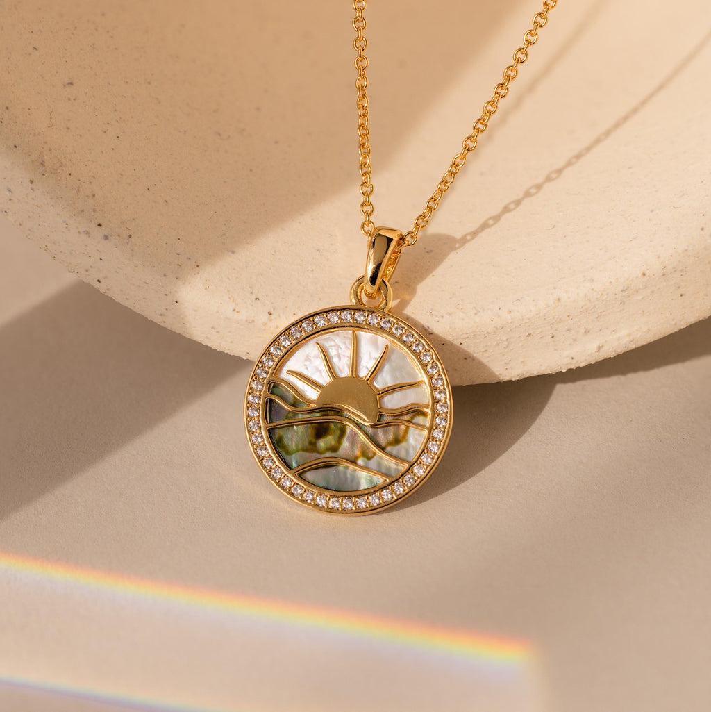 The Abalone Sunrise Pendant Necklace in 18K Gold features a round sun and wave design with small gems, beautifully displayed on a neutral background.