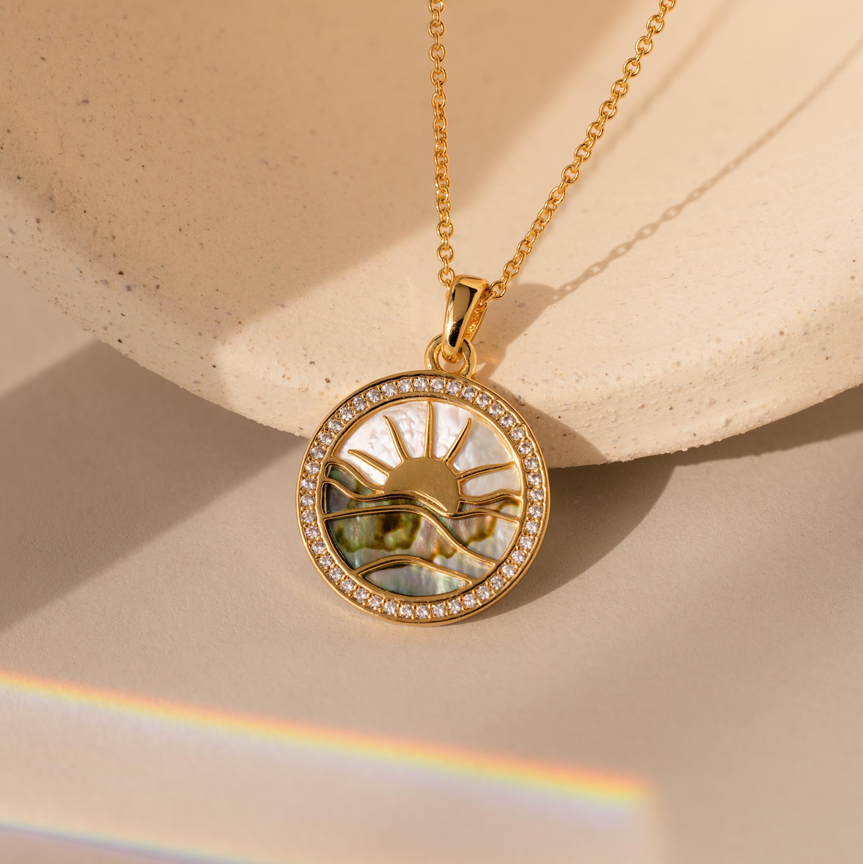 The Abalone Sunrise Pendant Necklace in 18K Gold features a round sun and wave design with small gems, beautifully displayed on a neutral background.