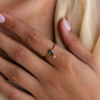 A hand with light pink nails wears the Duo Birthstone Drop Ring, showcasing a blue gemstone and a small clear stone in an elegant gold setting.
