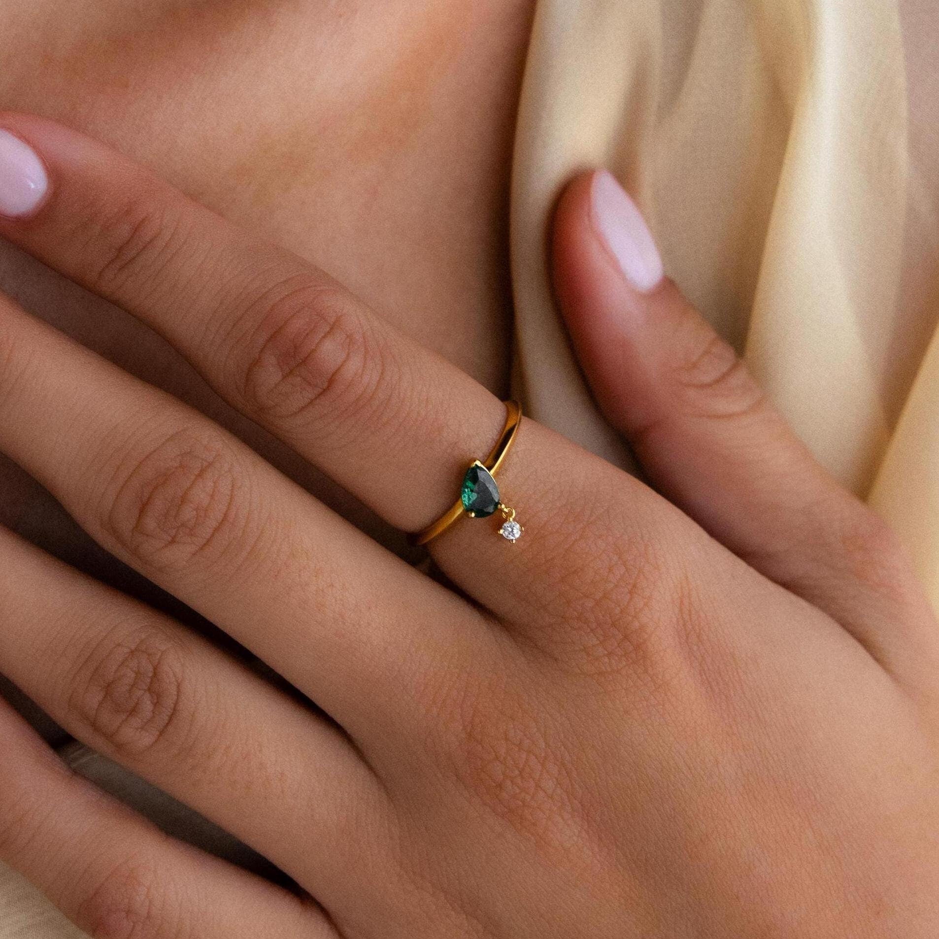 A hand with light pink nails wears the Duo Birthstone Drop Ring, showcasing a blue gemstone and a small clear stone in an elegant gold setting.