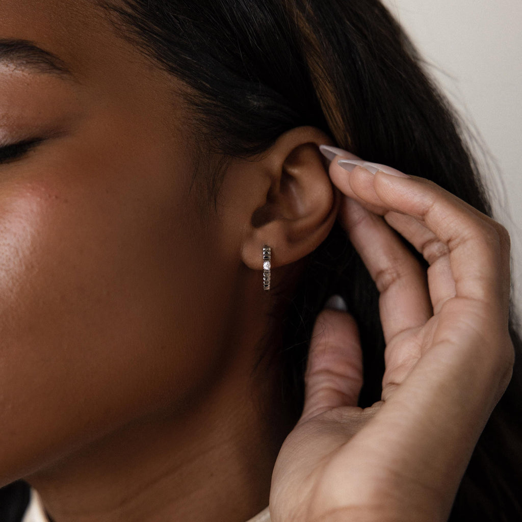 A close-up of a woman touching her ear, featuring the elegant Diamond Ribbed Huggies—dainty hoops with a ribbed design and sparkling stones.