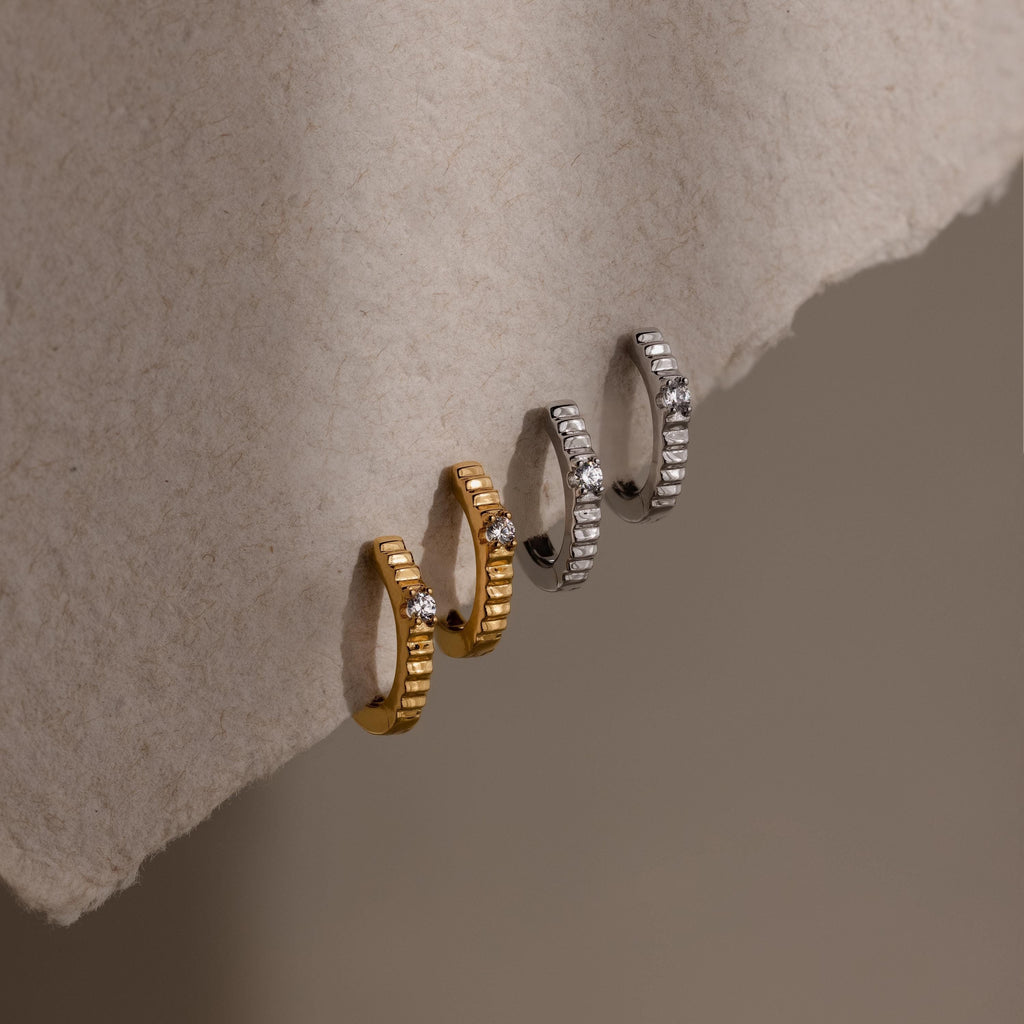 Two gold and one silver Diamond Ribbed Huggies, each with a ribbed design and diamond accents, are displayed on a soft beige textured surface.