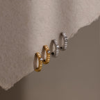 Two gold and one silver Diamond Ribbed Huggies, each with a ribbed design and diamond accents, are displayed on a soft beige textured surface.