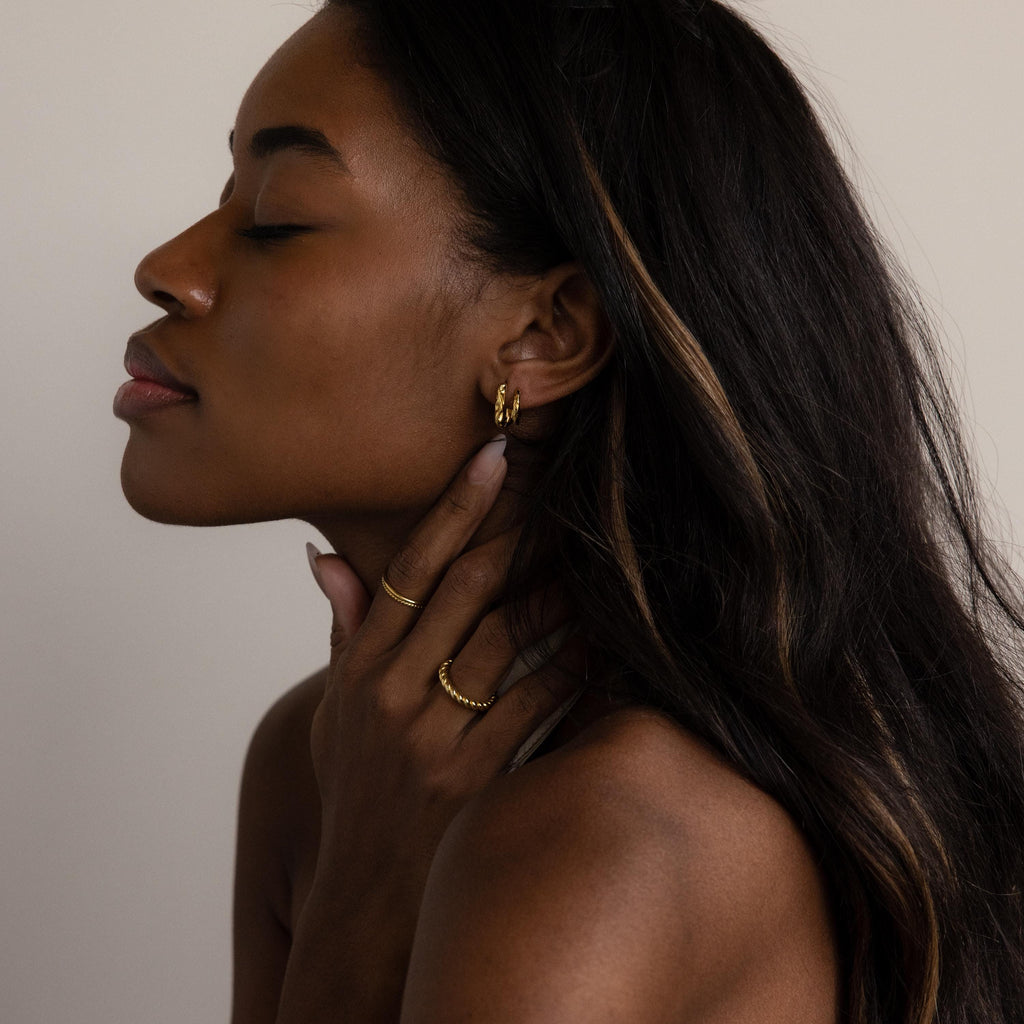 A woman with closed eyes touches her neck, wearing Hammered Rectangle Hoops and geometric rings, set against a neutral background.