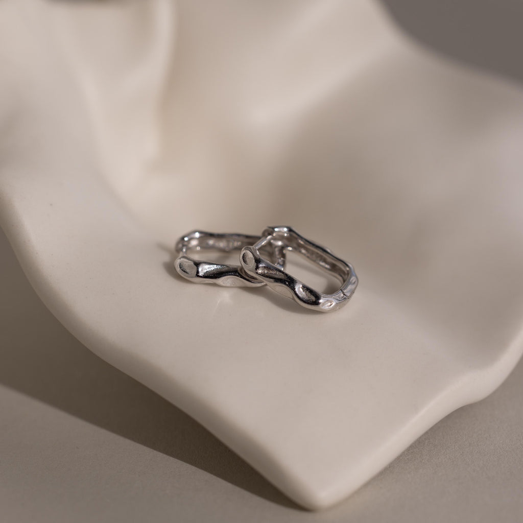 The Hammered Rectangle Hoops feature organic, textured silver shapes and are displayed on a smooth, light-colored sculptural surface.
