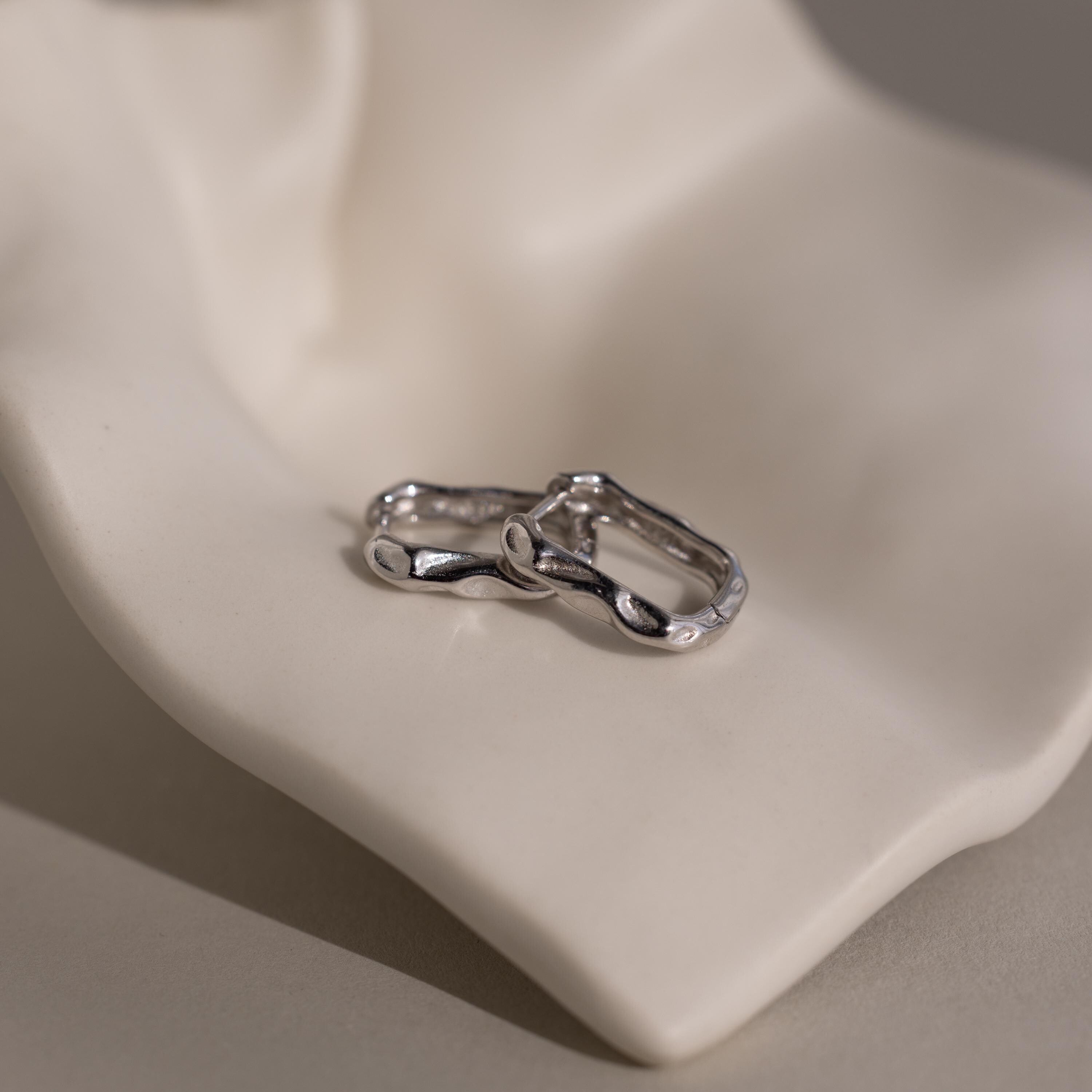 The Hammered Rectangle Hoops feature organic, textured silver shapes and are displayed on a smooth, light-colored sculptural surface.