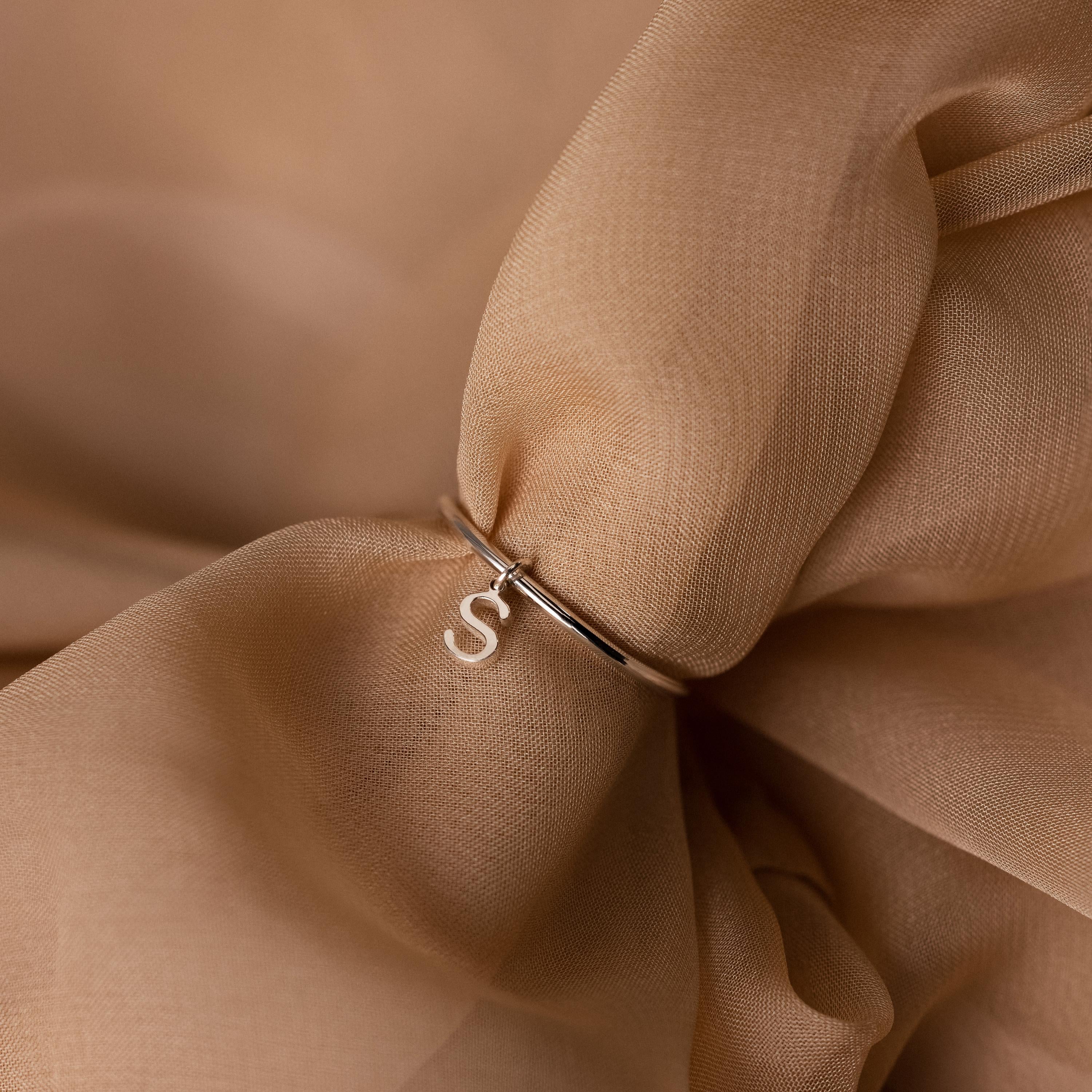 Close-up of the Dainty Initial Drop Ring, showcasing a silver S charm on soft beige fabric—a stylish choice for anyone who loves personalized jewelry.