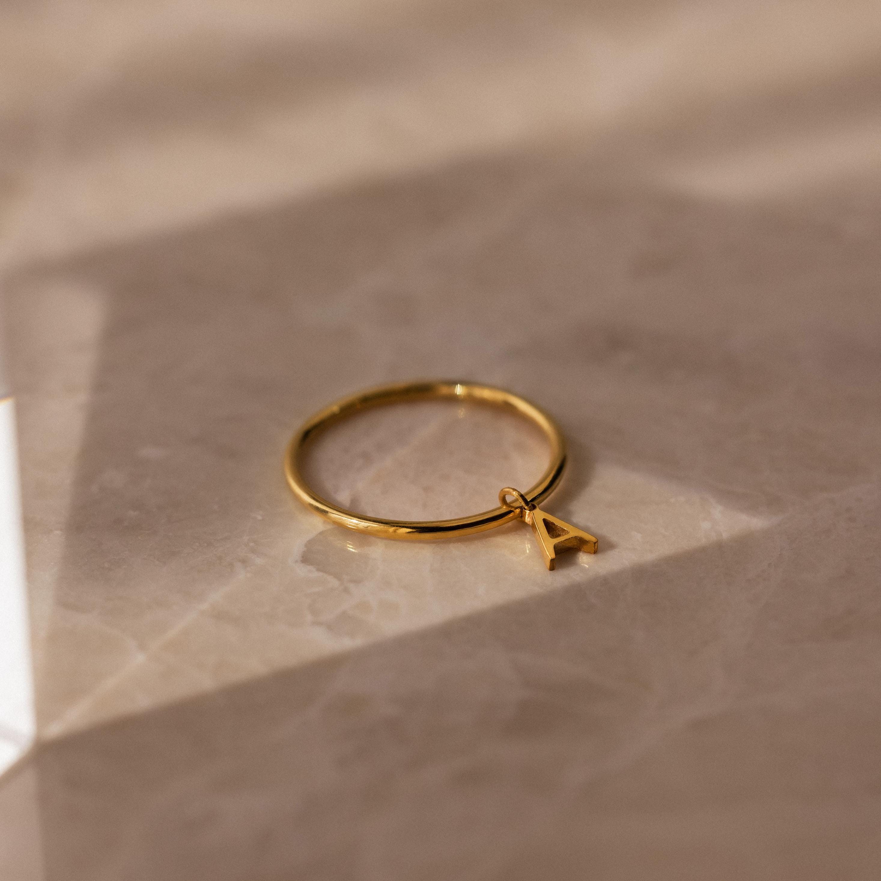 The Dainty Initial Drop Ring, adorned with a petite letter A charm, sits on beige marble in gentle sunlight—a subtle and personalized jewelry piece.