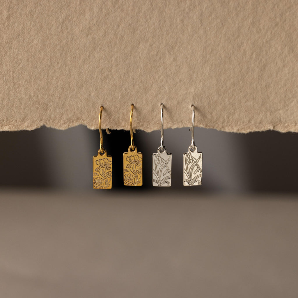 Two pairs of Rectangle Birth Flower Earrings, one gold and one silver, hang on a textured beige background—ideal floral jewelry for anyone who loves unique birth flower earrings.