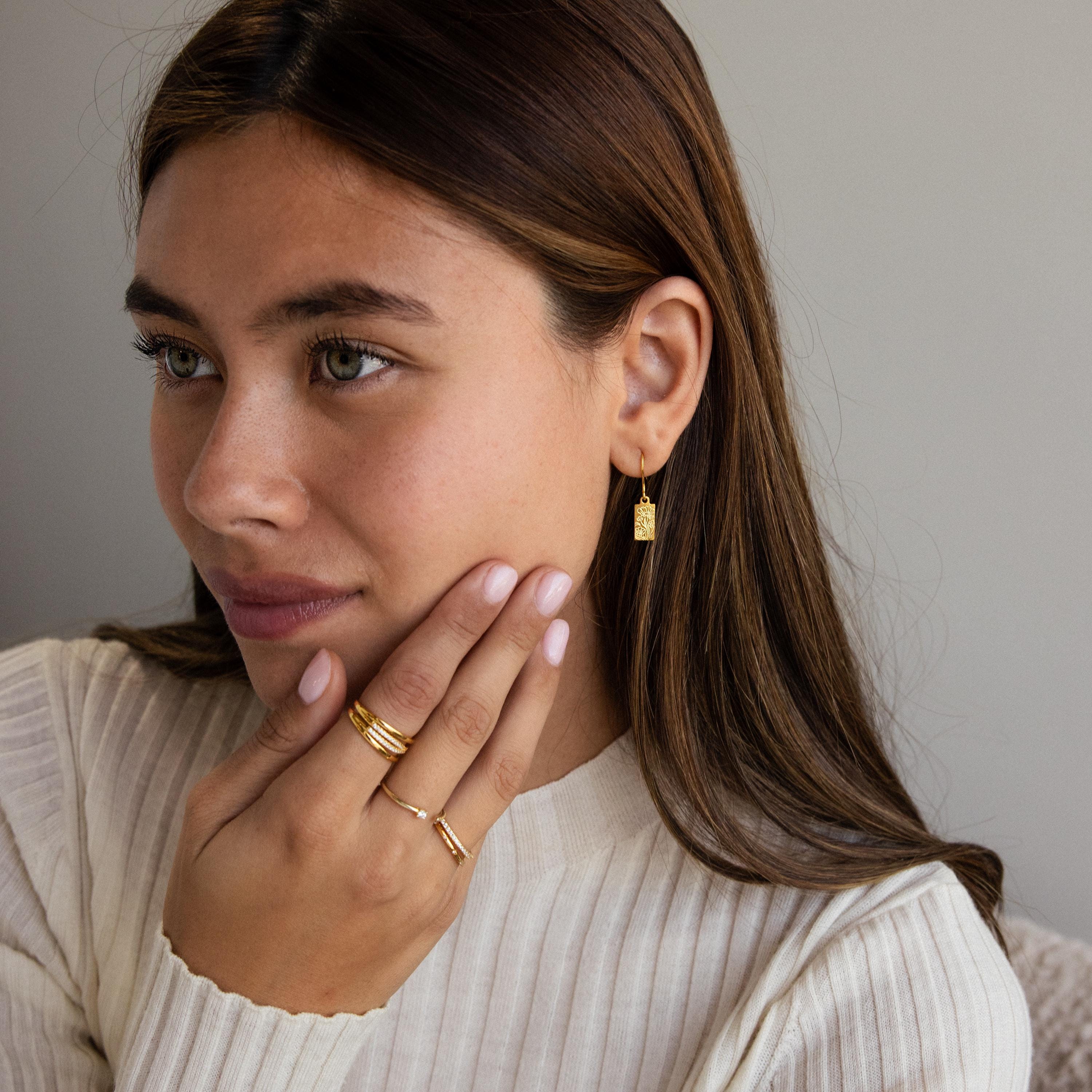 Woman with long brown hair wears Rectangle Birth Flower Earrings and rings, touches her face, and is dressed in a cream ribbed top.