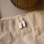 Rectangle Birth Flower Earrings with delicate leaf engravings are displayed on beige fabric next to a ribbed glass container.