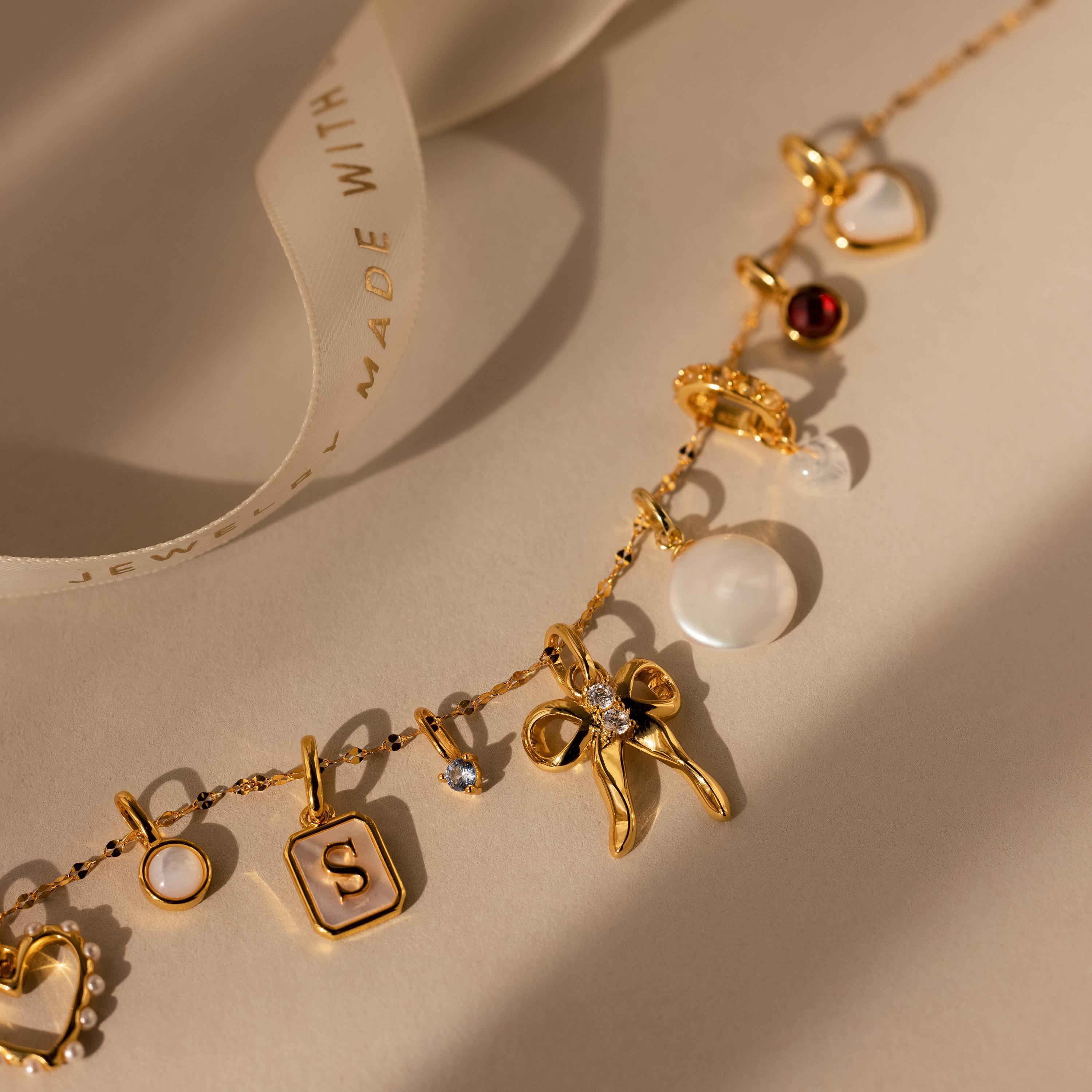 The Diamond Bow Charm necklace features assorted charms—pearl, heart, and letter S—making this coquette accessory stand out on a neutral background.