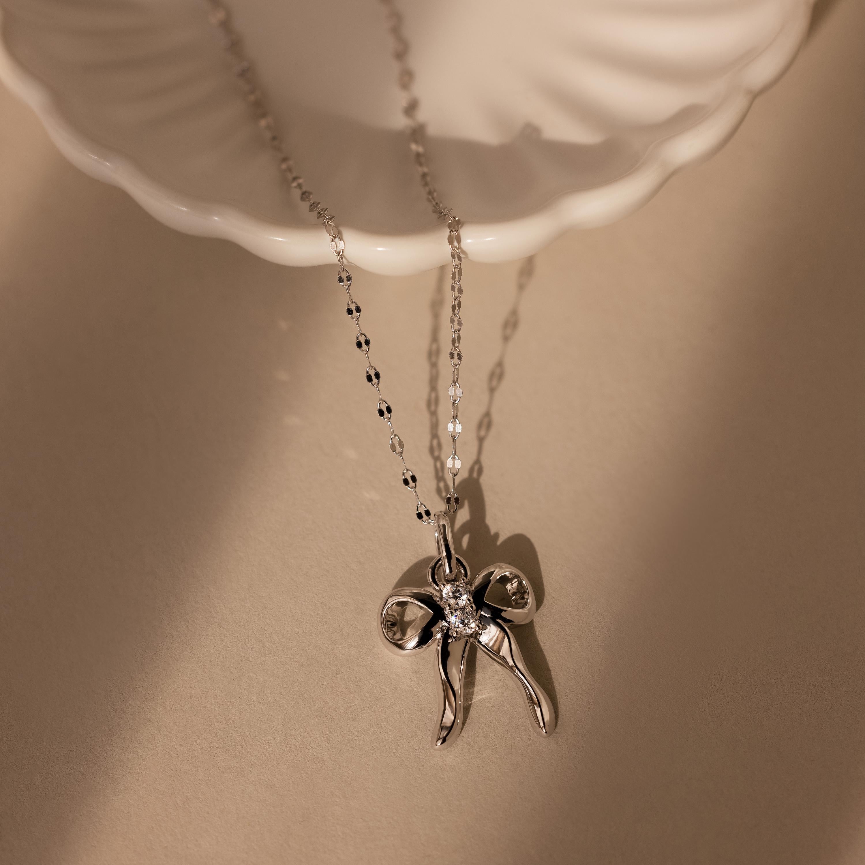 The Diamond Bow Charm in Sterling Silver features a bow-shaped pendant adorned with CZ diamonds and a central gem, elegantly displayed on a white dish under soft, warm light.