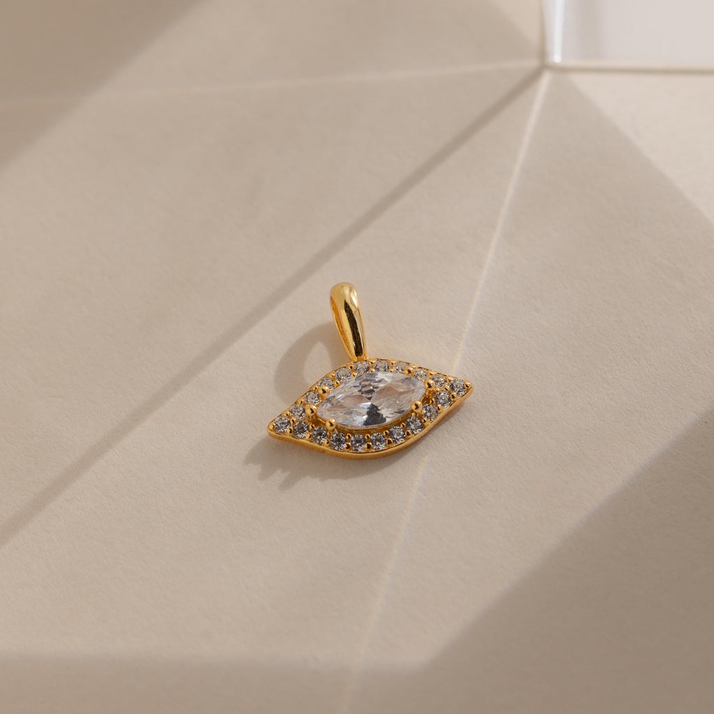 Gold Pave Evil Eye Charm on flat surface. The design features a marquise diamond in the center with pave gemstones surrounding it in the shape of an eye.