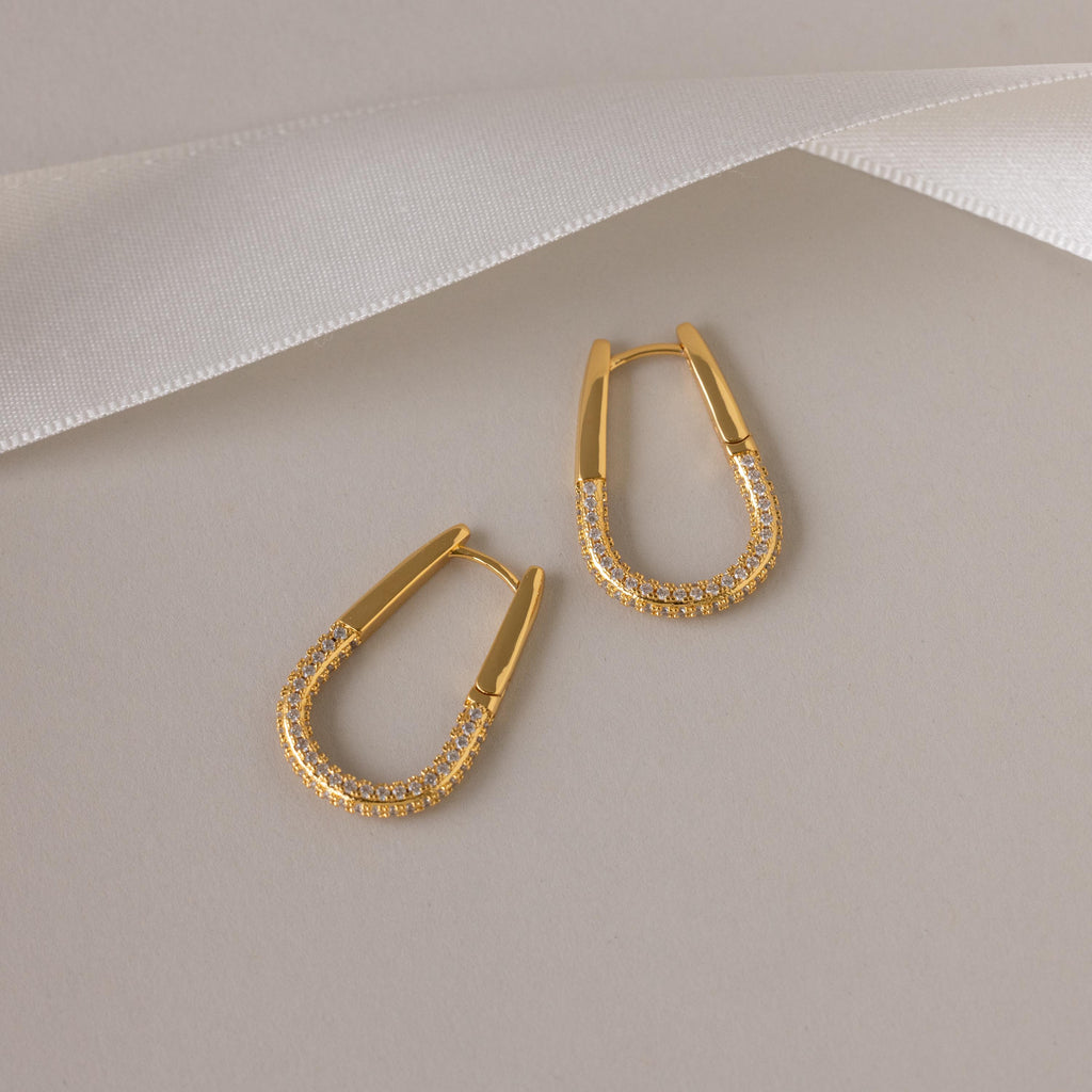 The Bold Pave Oval Hoops, featuring embedded pave diamonds, are displayed on a beige background with a white ribbon accent above.