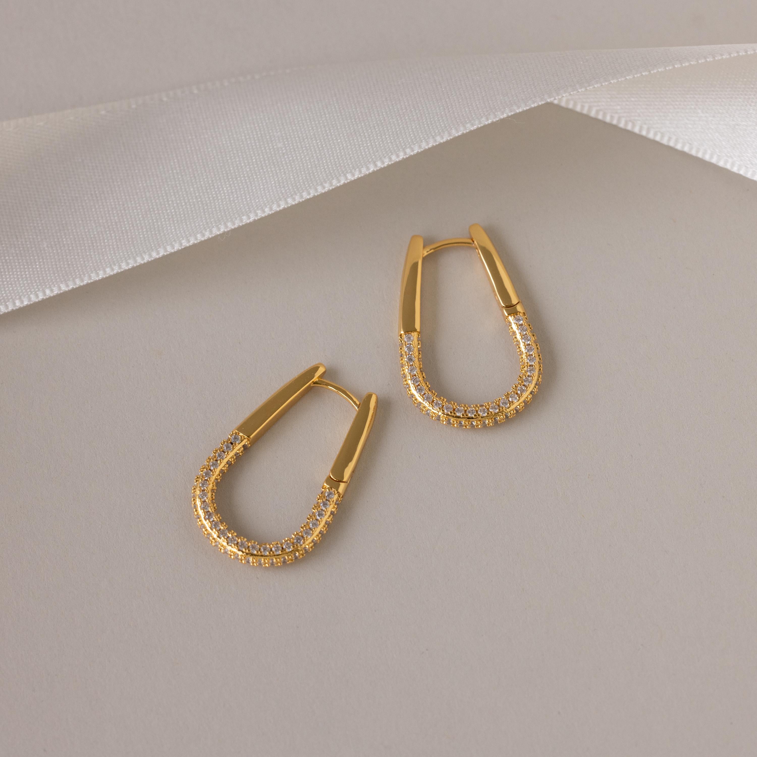 The Bold Pave Oval Hoops, featuring embedded pave diamonds, are displayed on a beige background with a white ribbon accent above.