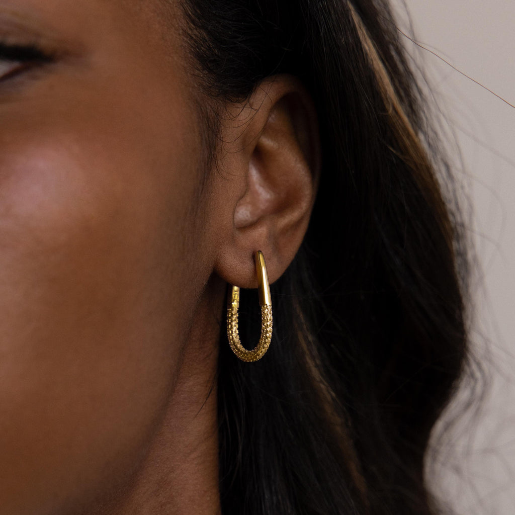 Close-up of a person's ear wearing the Bold Pave Oval Hoops—textured gold oval earrings with sparkling pave diamonds—partially framed by dark hair.