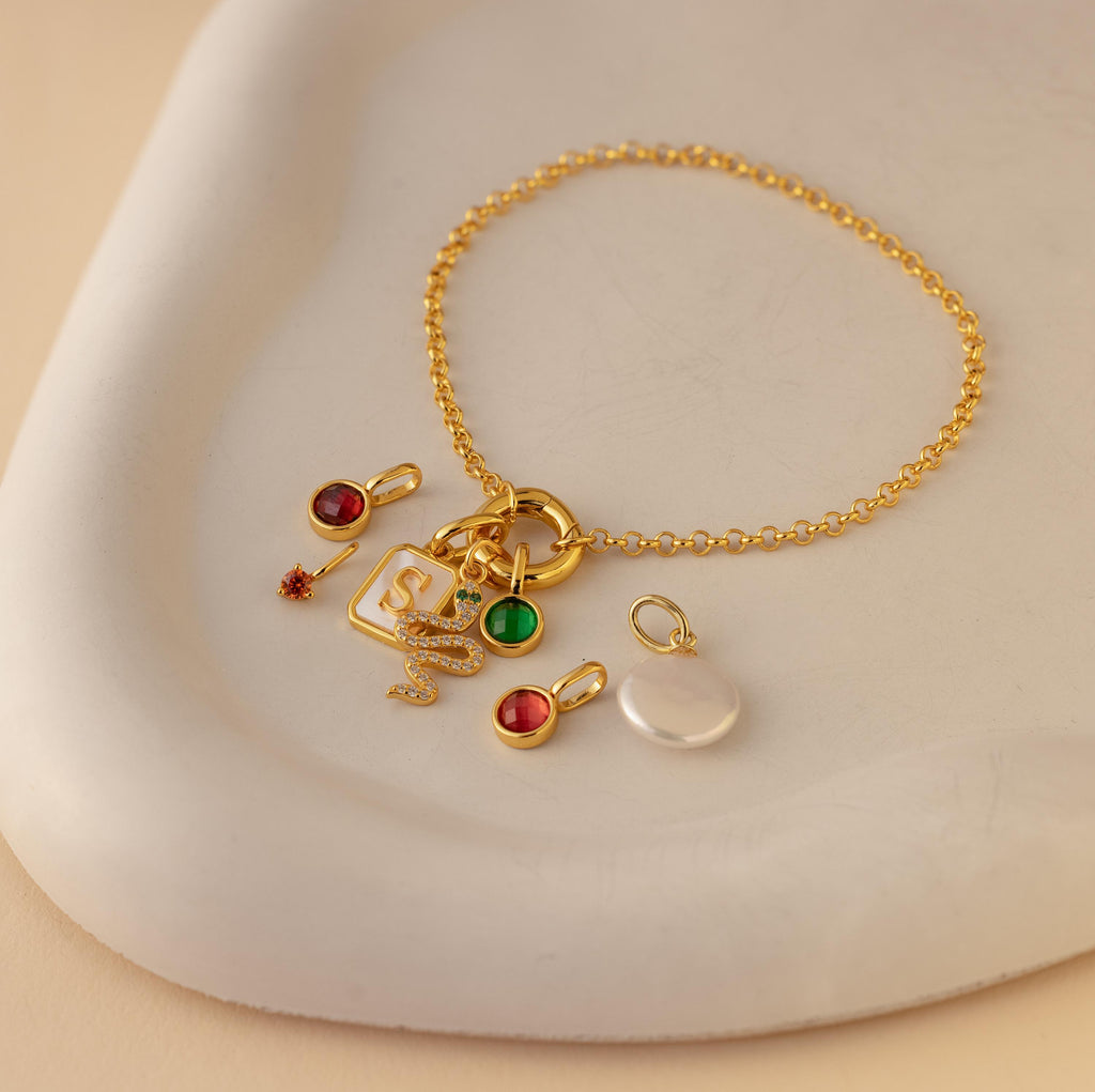 A gold rolo circle bracelet laying on a dish, adorned with different charms including a gold diamond snake charm