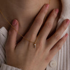 A hand touches the chest near a gold Pearl Diamond Drop Necklace with a small pendant, elegantly resting over a cream-colored sweater.