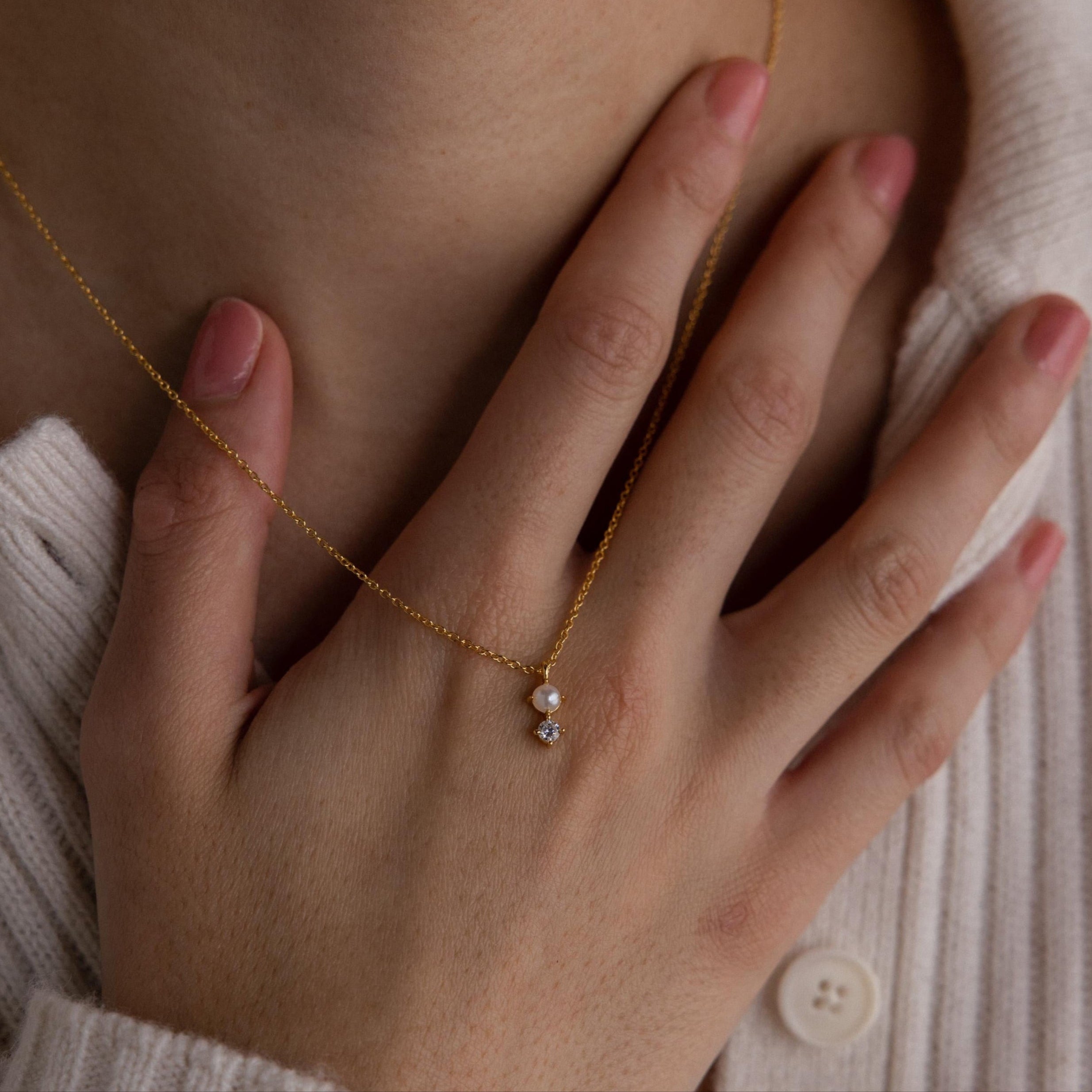 A hand touches the chest near a gold Pearl Diamond Drop Necklace with a small pendant, elegantly resting over a cream-colored sweater.