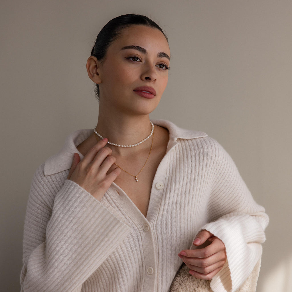 A woman in a cream ribbed sweater and layered necklaces, including a delicate Pearl Diamond Drop Necklace, gazes thoughtfully to the side against a plain background.