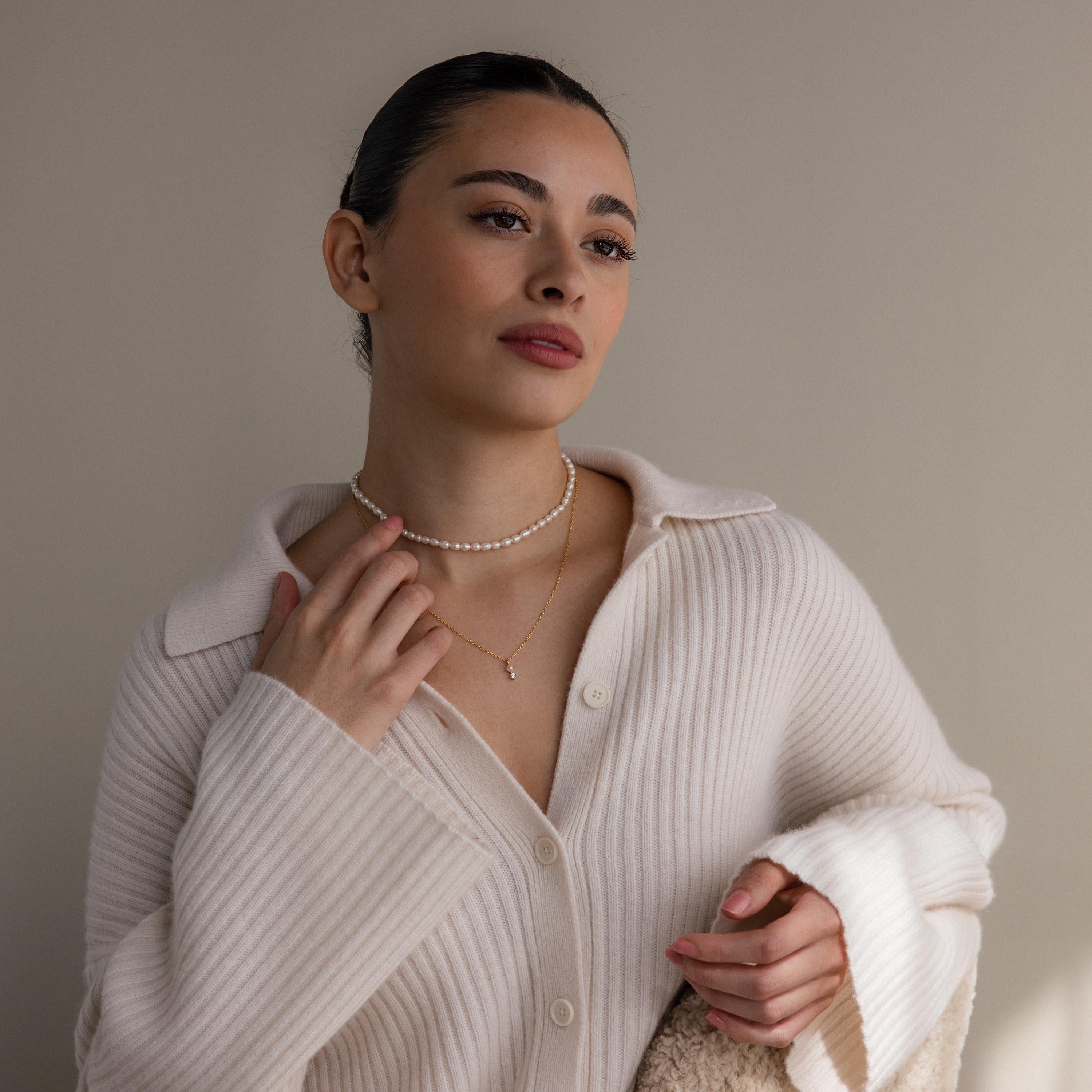 A woman in a cream ribbed sweater and layered necklaces, including a delicate Pearl Diamond Drop Necklace, gazes thoughtfully to the side against a plain background.