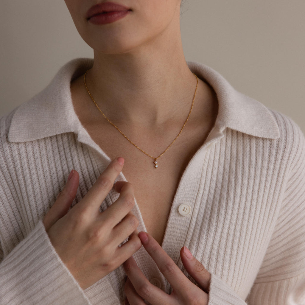 A woman in a cream ribbed shirt touches her collar, showcasing the Pearl Diamond Drop Necklace—a gold necklace with a delicate pearl diamond pendant.