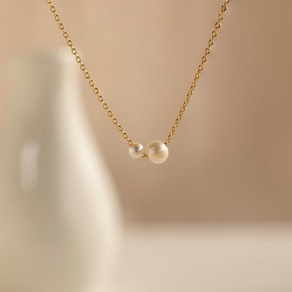 The Dainty Duo Pearl Necklace features a gold chain with two pearls, ideal as dainty jewelry or a thoughtful wedding gift. Displayed against a blurred white vase and beige background.