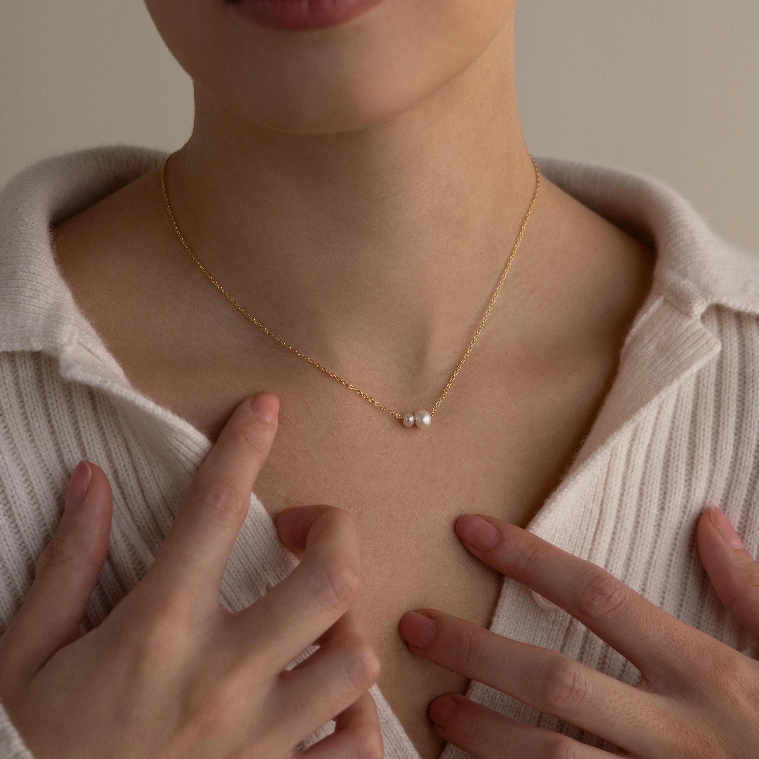 A person wears the Dainty Duo Pearl Necklace featuring two pearls resting at the collarbone over a cream ribbed top—an elegant wedding gift choice.