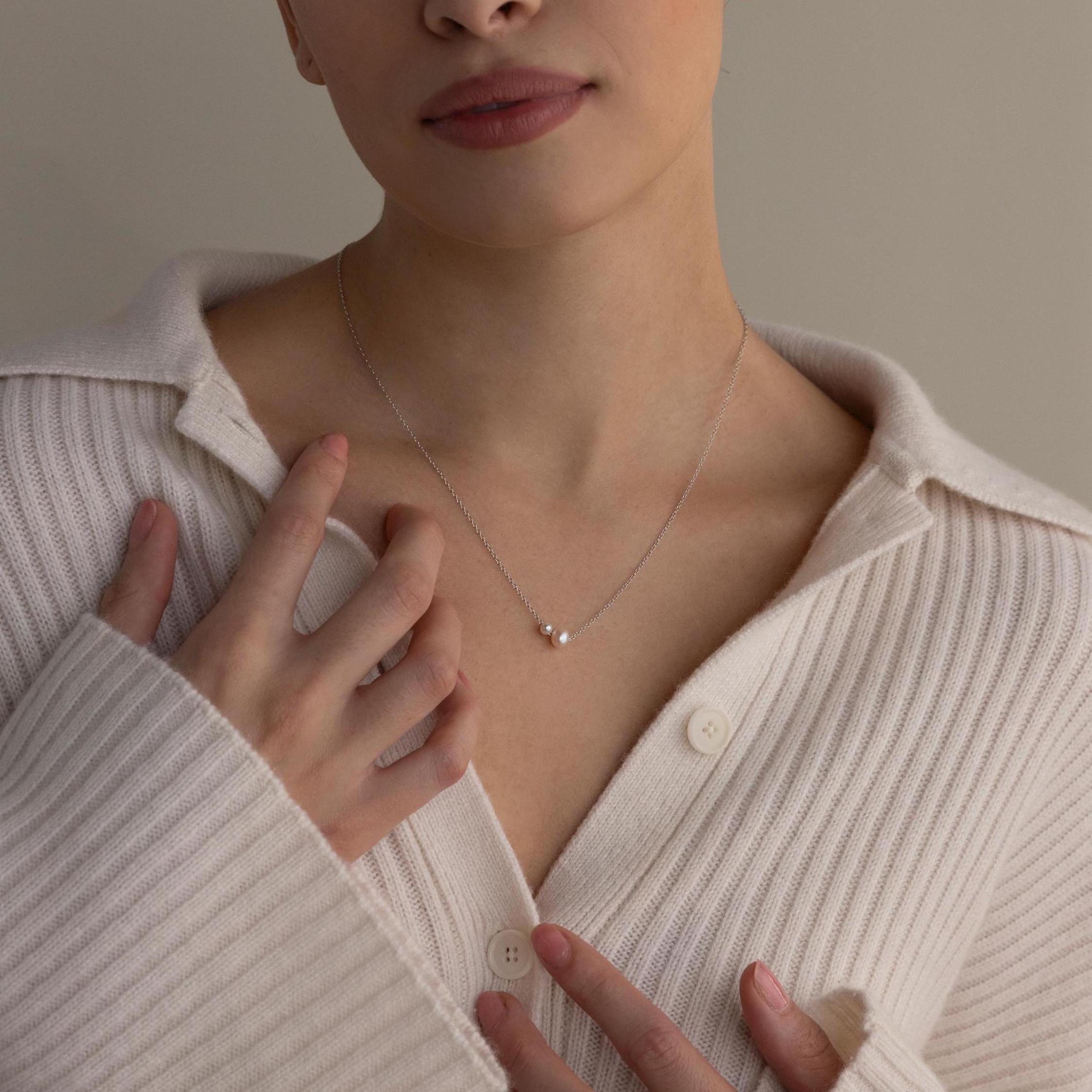 A woman in a cream sweater touches the Dainty Duo Pearl Necklace, featuring a delicate pearl and small pendant around her neck.