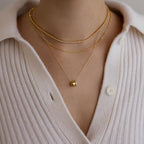 Close-up of a person wearing elegant jewelry: three gold necklaces, including the dainty Pave Duo Circle Necklace, paired with a cream-colored button-up knit top.