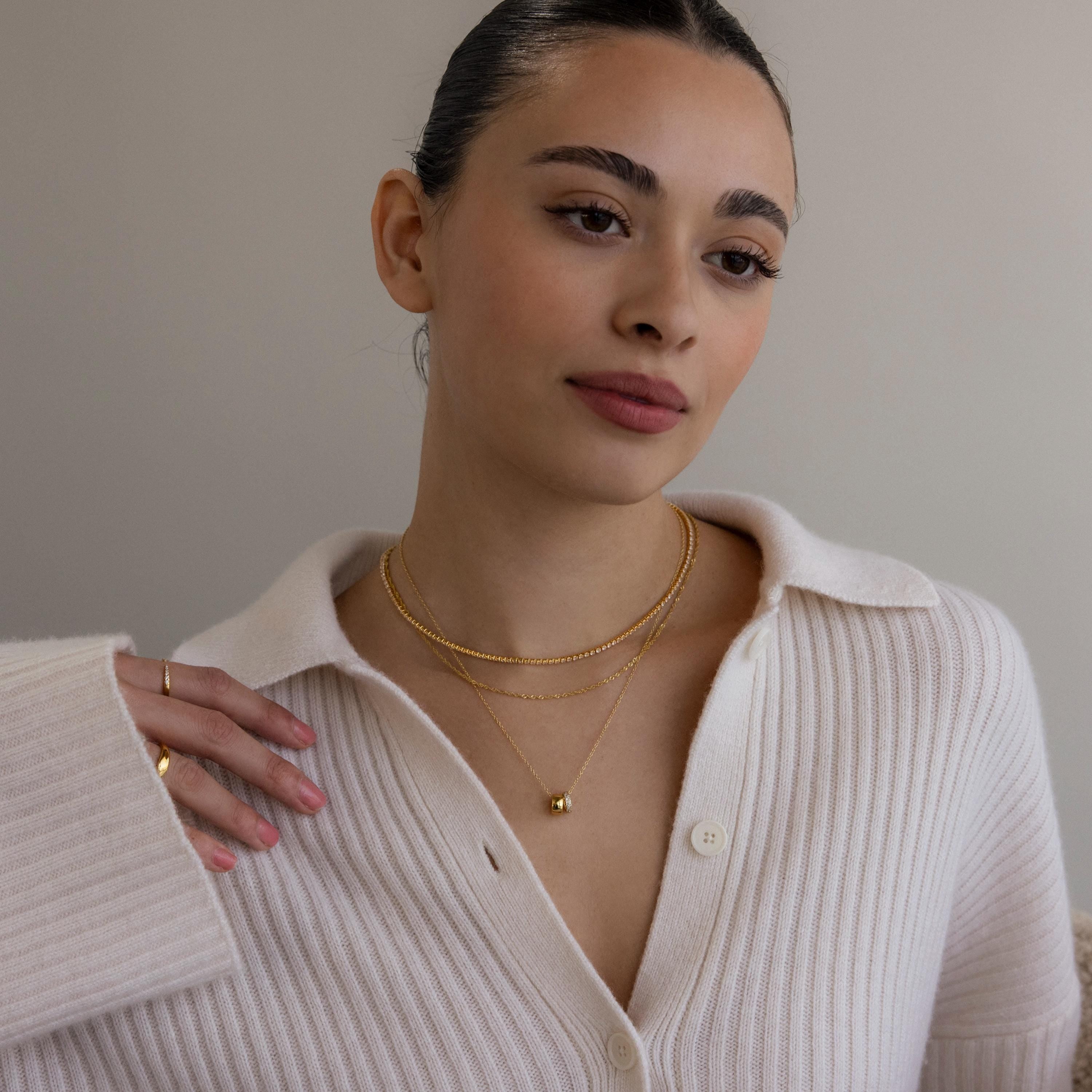 Woman in a cream ribbed sweater wearing elegant jewelry, including the Pave Duo Circle Necklace and layered gold rings, looking serene against a neutral background.