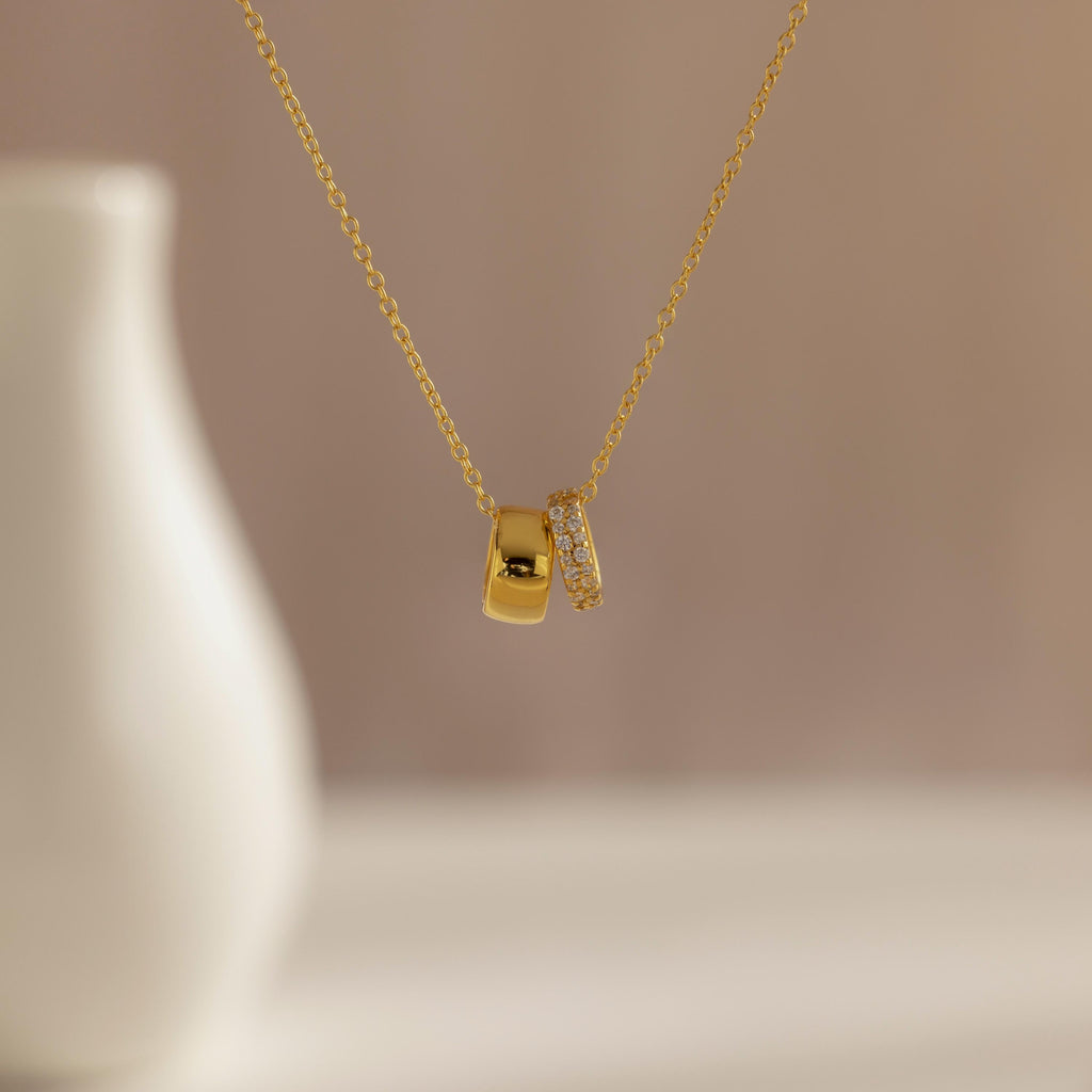 The Pave Duo Circle Necklace features two pendants—one plain, one with pavé stones—for a dainty, elegant look, shown beautifully in front of a blurred vase.