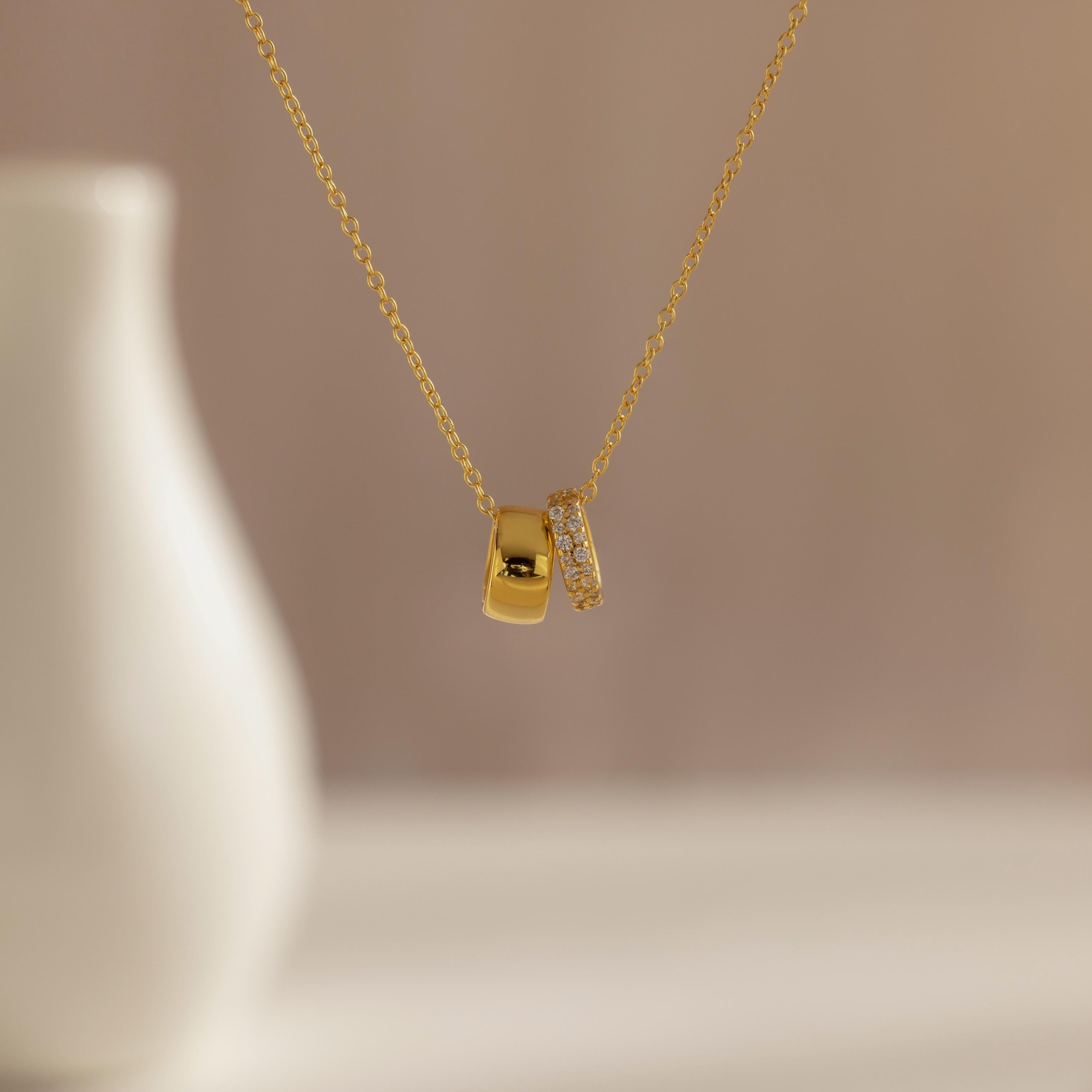 The Pave Duo Circle Necklace features two pendants—one plain, one with pavé stones—for a dainty, elegant look, shown beautifully in front of a blurred vase.